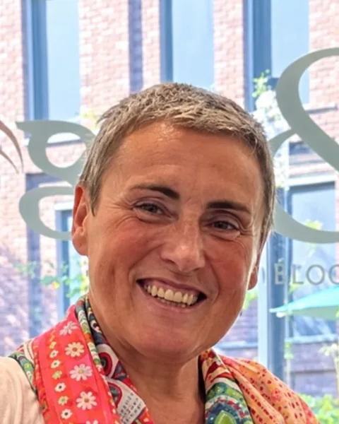 Portret van Greta Fiers, met kort grijs haar en een kleurrijke sjaal, lachend naar de camera.