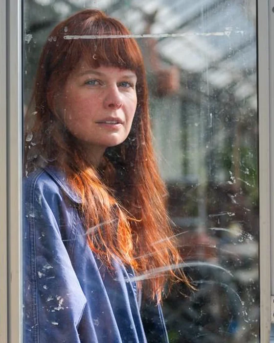 Jozefien Mombaerts, met lang rood haar en een blauwe werkjas, kijkt vanachter een glazen deur van een serre naar de camera.